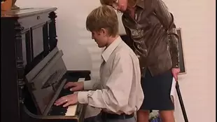 A mature European woman in spectacles takes control of her young piano instructor